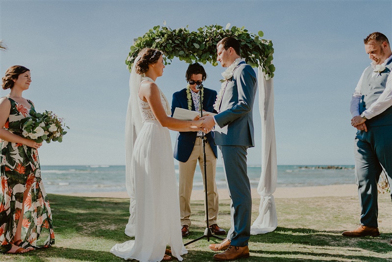 A bride and groom stand facing one another as they hold hands during their civil wedding ceremony. The bride is in a simple floor length white wedding dress with a long view and her hair tied into a dressy bun hairstyle. The groom is in a blue suit with brown dress shoes. Between the two you can see their officiant who wears beige slacks and a navy blue suit jacket and has a lea around his neck. The officiant is holding his speech. A bridesmaid in a floral floor length dress stands to the left of the bride and groom holding a bouquet and a groomsman stands to the right with his hands folded in front, and wearing blue dress pants and vest with a white shirt and brown shoes | Hawaii Wedding Photographer, Hawaii Elopement Photographer, Hawaii Elopement Inspiration | chelseaabril.com