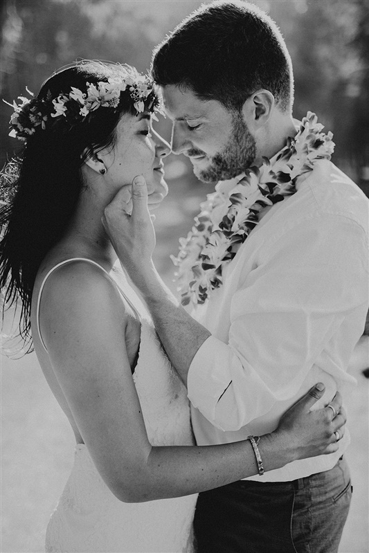 A man and a woman embrace one another. The woman has her arms wrapped around the man's waist and he is cupping her chin with his left hand. The woman is in a simple tight fitting white dress and a flower crown sits on her dark hair. The ma is in dress pants and a white dress shirt that is rolled up to the elbows. He is wearing a lea. The photo is in black and white and it Is of a non traditional wedding couple | Hawaii Wedding Photographer, Hawaii Elopement Photographer, Hawaii Elopement Inspiration | chelseaabril.com