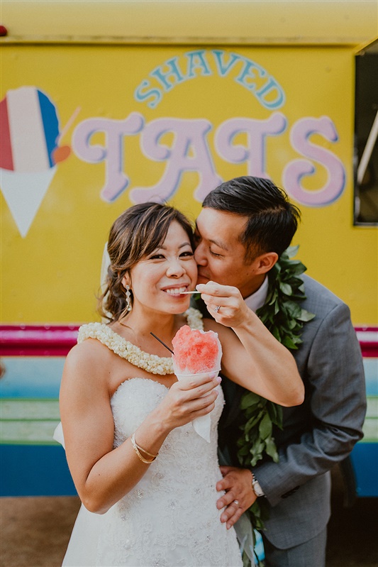 A bride and groom stand outside a bright yellow shaved ice truck as they eat some flavoured shaved ice in a paper cone . the man has leaf lea around his neck, the bride is in a white wedding dress with her brown hair pulled back, The groom is in a grey suit | Hawaii Wedding Photographer, Hawaii Elopement Photographer, Hawaii Elopement Inspiration | chelseaabril.com