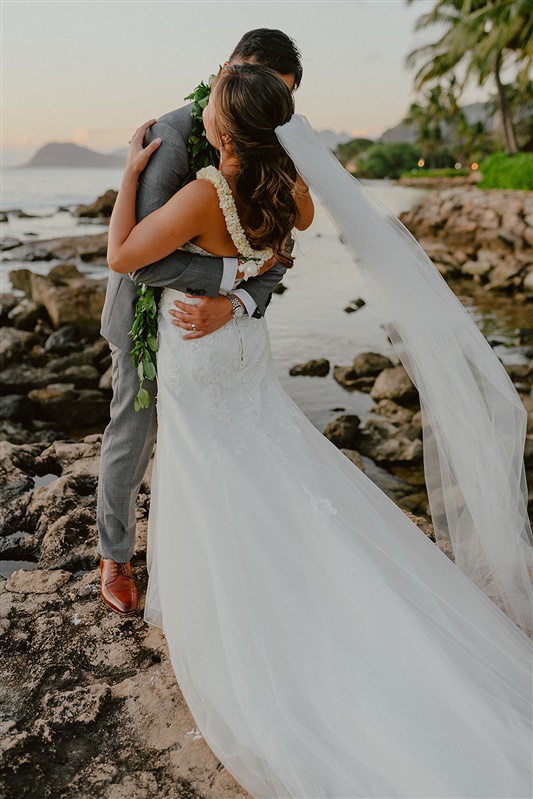 A bride and groom embrace in a hug following their civil wedding ceremony. The bride is wearing a  white wedding dress and a long floor-length veil. The groom is in a light grey suit and jacket with brown shoes and a leaf lea around his neck. They are standing on a shoreline | Hawaii Wedding Photographer, Hawaii Elopement Photographer, Hawaii Elopement Inspiration | chelseaabril.com