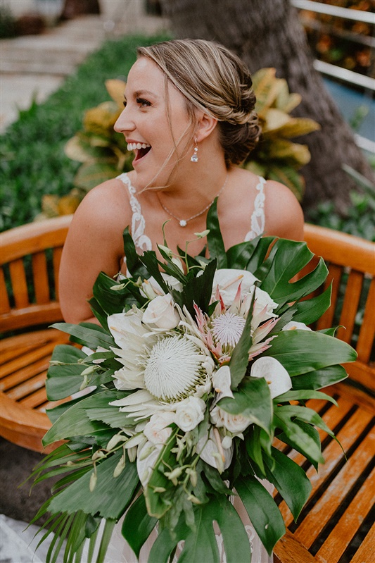 A bride smiles as she poses with her large bouquet featuring tropical flowers and greenery outside her Cafe Julia Wedding