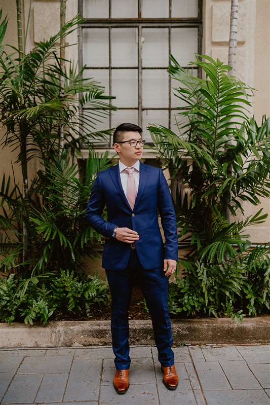 A groom stands in front of a building with lots of dark and tropical greenery. He's wearing a dark blue dress pants and jacket, pink tie, white shirt and brown dress shoes and black glasses.