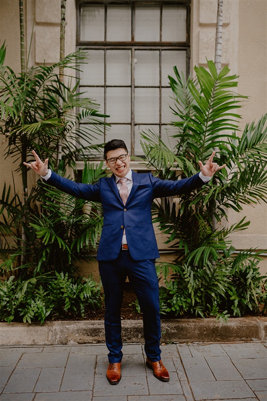 A groom grins sillily while throwing up some devil horns with his hands. He is wearing a dark blue suit jacket and pants, a white dress shirt, pink tie and brown shoes and black glasses