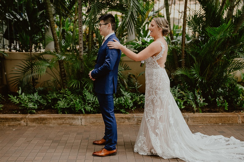 A bride places her hand on her groom's shoulder as she approaches him from behind. She is wearing a detailed floor-length white wedding dress. The groom stands waiting to get a first look at his bride. He is wearing a blue suit with brown dress shoes.
