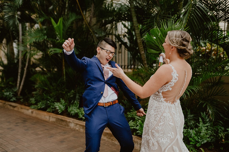 A groom dances with excitement as he sees his bride for the first time. He is wearing a blue suit with a brown belt, white dress shirt and pink tie. The bride is in a detailed white backless wedding dress.