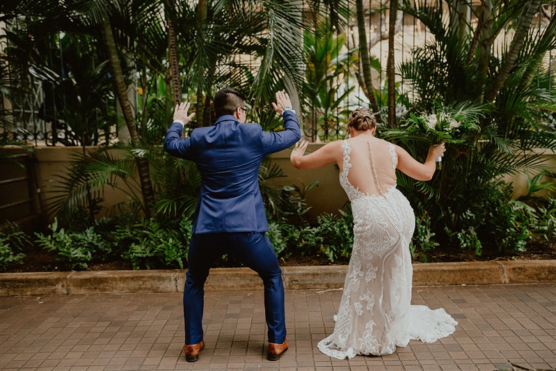 A bride and groom throw their hands in the air and wiggle their bums at the camera as they dance sillily. The bride is in a detailed white wedding dress and the groom is in a blue suit and brown dress shoes. They both are standing outside of Cafe Julia