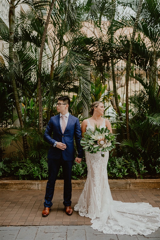 A bride and groom stand outside at their Julia Cafe wedding. They stand side by side but are looking in opposite directions. The groom is in a blue suit, white dress shirt, pink tie, and brown dress shoes. the bride is in a detailed white wedding dress with a semi long train and is holding a large bouquet of tropical greenery and flowers.