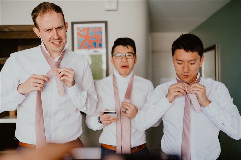 A groom stands with a phone in his hand and a groomsman on each side of him. All three men have a silly look on their face as they attempt and fail to tie their neck ties. 