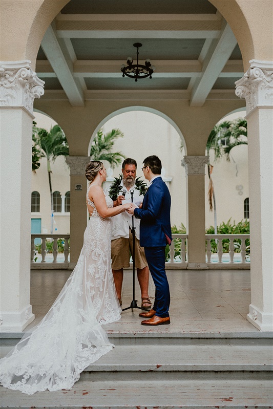A bride and groom stand facing one another hand in hand at their Cafe Julia wedding in Honolulu, Hawaii. An Officiant can be seen reading his speech between the two. 