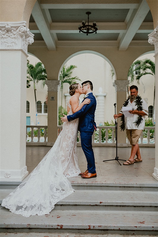 A bride in a detailed white wedding dress with a long train and a groom in a blue suit and brown dress shoes share their first kiss after they become married.