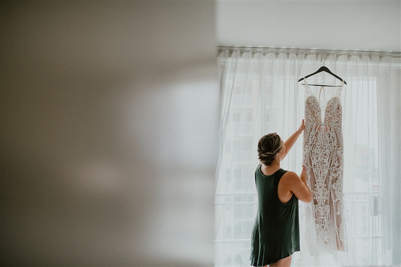 A bride inspects her white wedding dress which is hanging in the light of a window