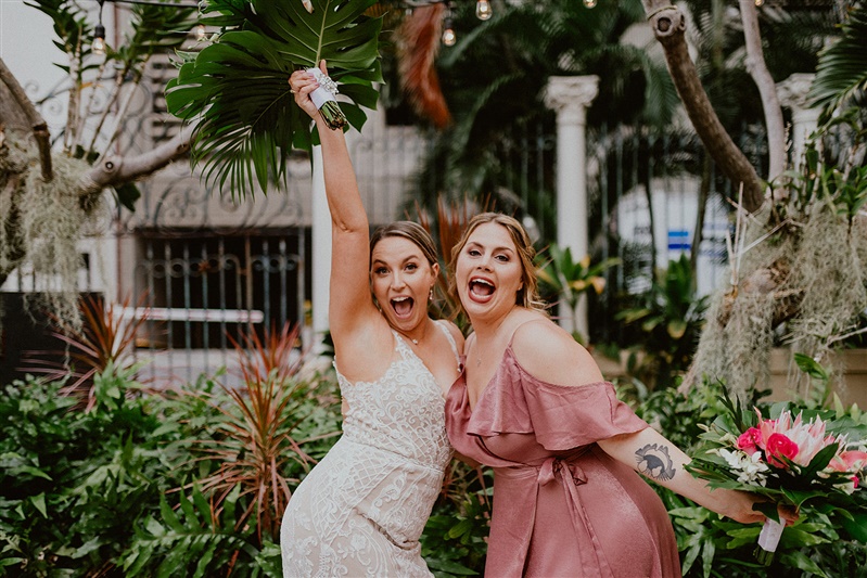 A bride and her bridesmaid celebrate outside their cafe Julia wedding. the bridesmaid is in a pink formal dress and the bride in a white detailed wedding dres.