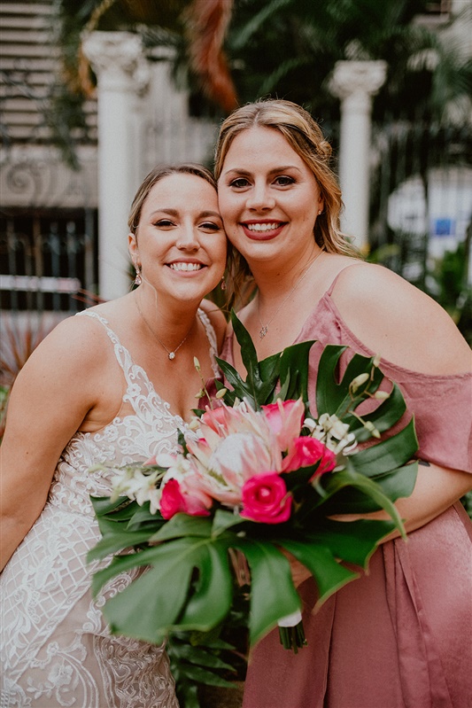 A bride poses smiling with her bridesmaid at a Cafe Julia Wedding. The bride is wearing a detailed white dress and simple necklace and earrings. Her blonde hair is pulled back into a bun. The bridesmaid is wearing a pink dress and is holding a large bouquet of tropical flowers and greenery,