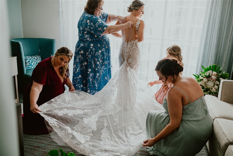 A bride stands with her back to the camera while wearing a detailed white wedding dress. Her mother stands behind her buttoning up the back while three bridesmaids help spread out the dress's train