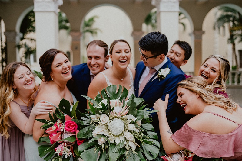 A bride and groom cuddle together in a group laughing with their bridesmaids and groomsmen. All are dressed in formal wear and the bridesmaids are holding large tropical bouquets.