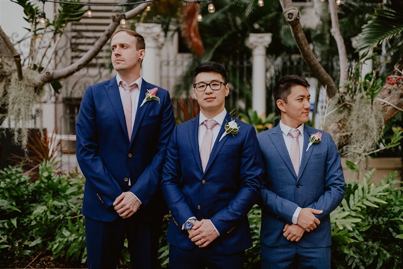A groom stands with his groomsmen on either side of him. They all wear dark blue suits, white shirts and pink ties. Each has a calla lily boutonniere.