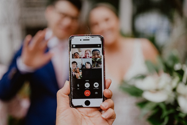 A cell phone is shown in the middle of the photo with a video chat with several people on the screen. A bride and groom are out of focus and align from the background to all the people in the video.