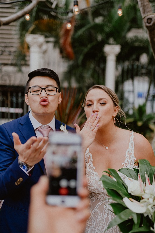 A bride and groom blow a kiss to wedding attendants who are participating through a video chat on a cell phone. The cell phone is blurred in the foreground. 
