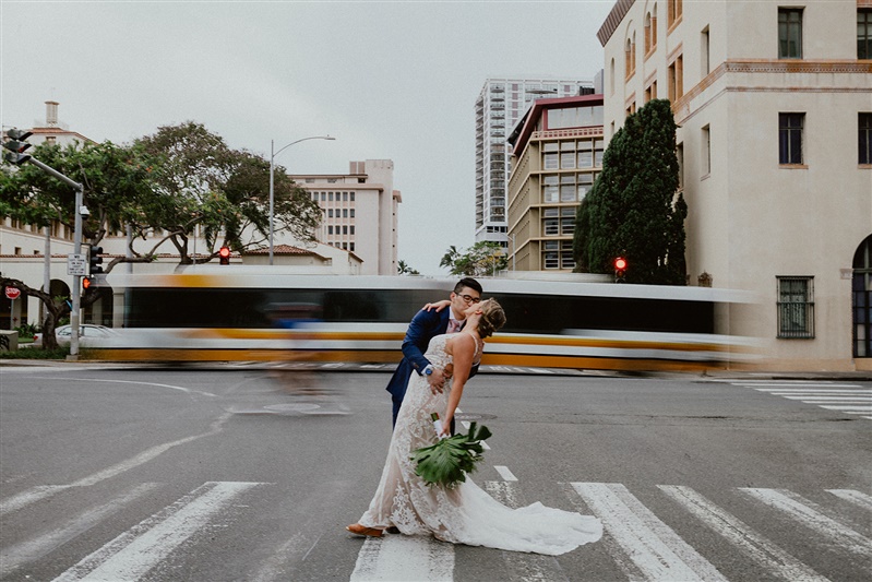 A bride and groom kiss in the middle of s street crosswalk. A bus goes by in a blue in the background.