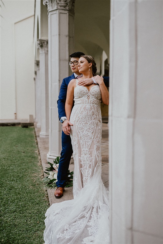 A bride and groom pose for a portrait together at Cafe Julia in Honolulu. The bride is in a long detailed white dress and the groom is in a blue suit with brown dress shoes. 
