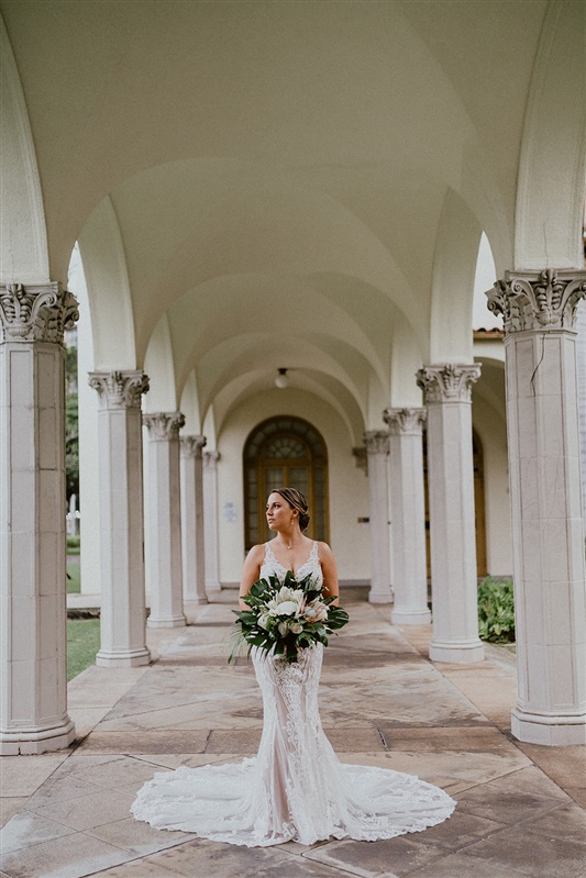 A bride stands between a column of pillars. She wears a long white detailed wedding dress and holds a large bouquet of tropical flowers.