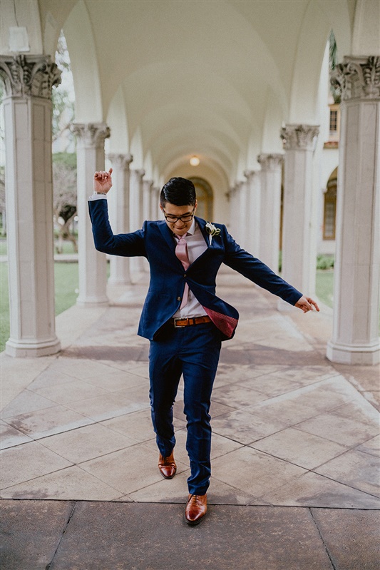 A groom dances in front of the camera. He is in a blue suit, white shirt and pink tie with brown dress shoes.