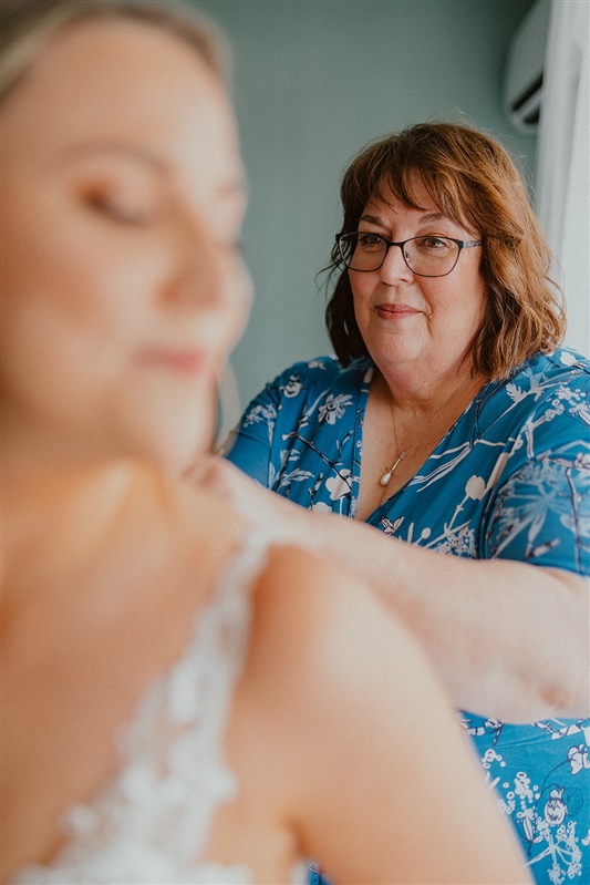 A mother buttons up a. bride's dress. The bride is out of focus.