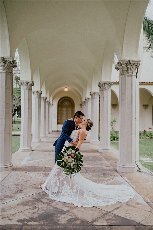 A groom tips back his bride and kisses her between a column of pillars. nThe bride is in a detailed white wedding dress, and is hold a large bouquet of tropical greenery and flowers. The groom is wearing a dark blue suit and pink tie.