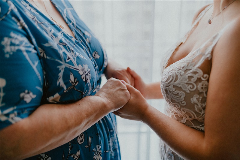 A mother and bride are seen from their shoulders to their waist, facing one another and holding hands.