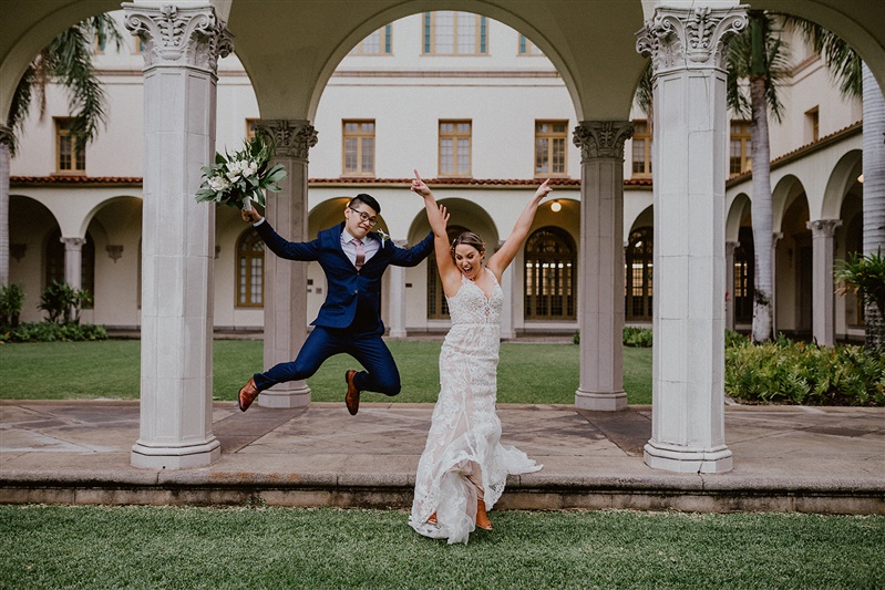 A bride and groom jump in the air for a fun posed photo together after their wedding. The bride is in a detailed white dress and the groom is in a blue suit with brown dress shoes. 