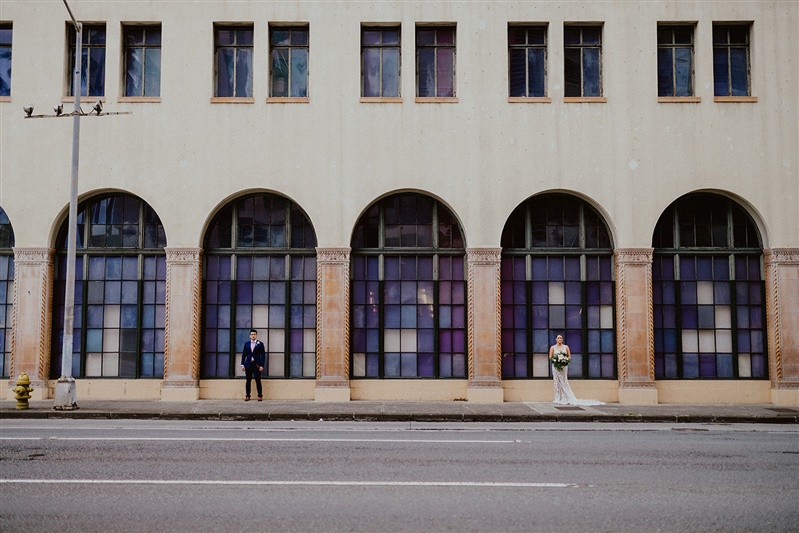A bride and groom stand a few meters away from one another and are photographed from across the street. The empty street is before them and behind them is an old building with large stained glass windows.
