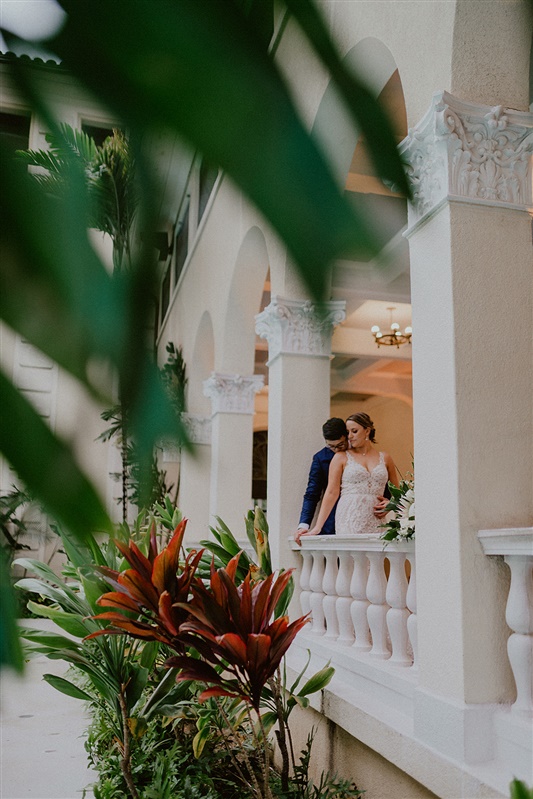 A bride and groom stand by a white railing with tropical plants surrounding theta a Hawaii hotel