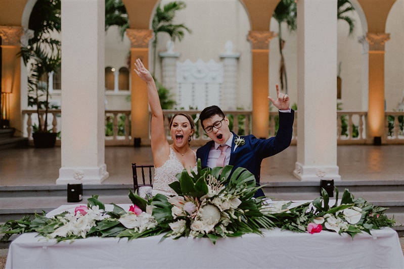 A bride and groom sit cheering at the heatable of their Cafe Julia wedding in Hawaii. Their table is decorated with lots of tropical flowers and greenery.