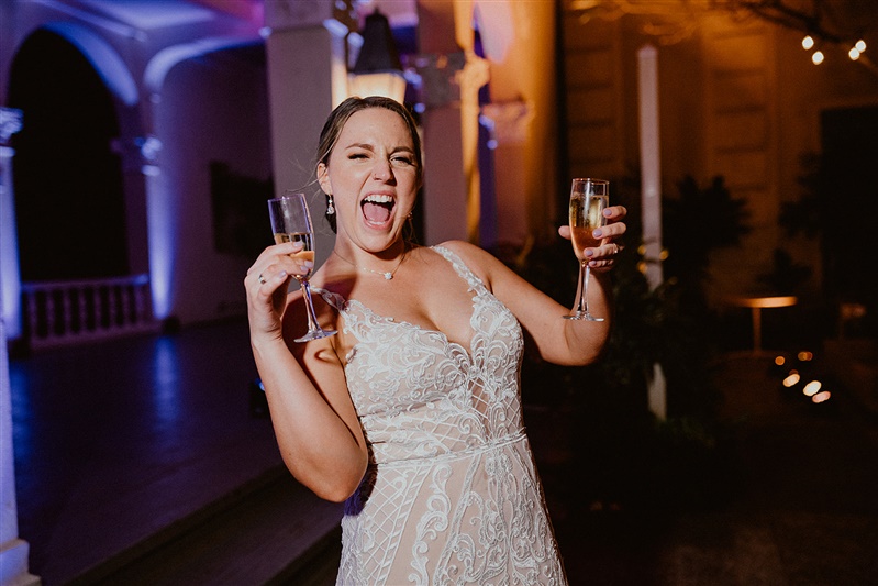 A bride dances with a glass of champaign in both hands. She is wearing a detailed white wedding dress.