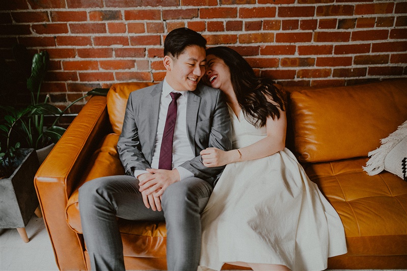 A bride and groom sit on a couch in the Seattle courthouse before their wedding ceremony. The couple are cuddled closely with the bride's chin on the groom's shoulder as the two laugh. The bride is wearing a simple but modern wedding dress and the groom is in a full grey suit with a Burgundy tie. 