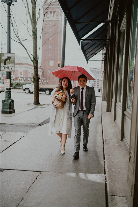 A man and woman walk down a Seattle sidewalk in the rain while holding an umbrella. The man is in grey suit and tie while the woman is in a white dress and formal coat and is holding flowers. They are both  on their way to their courthouse wedding. 