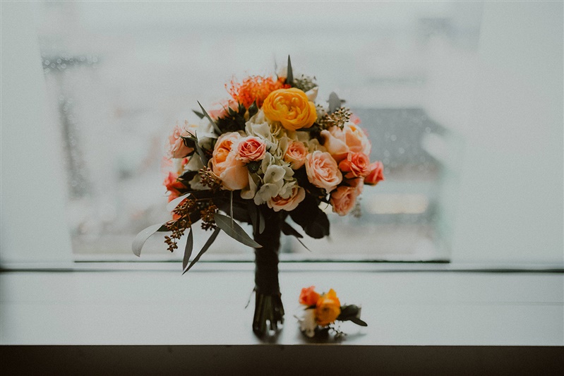 A bridal bouquet and boutonniere sit on a ledge against a rainy window pane in the Seattle courthouse. The flowers are light pinks, oranges and blush shades.