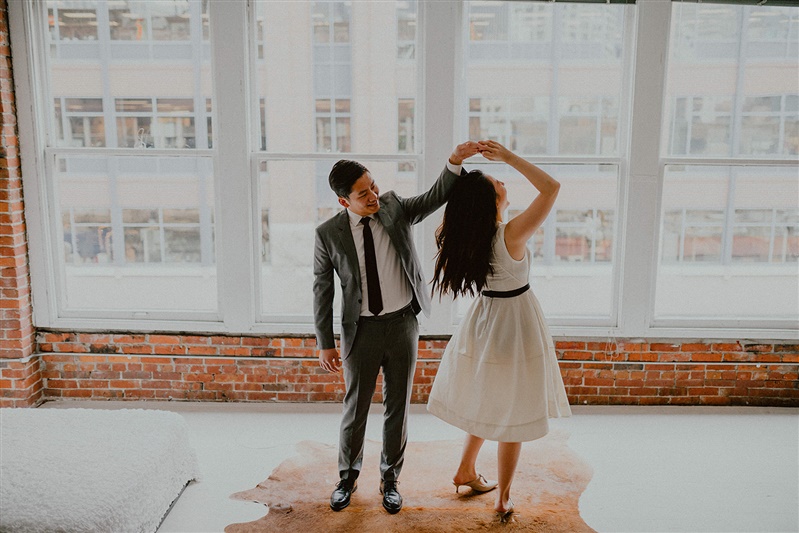 A bride and groom dance together in the Seattle courthouse before their wedding ceremony. The groom is spinning the bride while they both stand on a animal skin rug. The bride is wearing a white dress with black belt and the groom is in a full grey suit with a Burgundy tie.