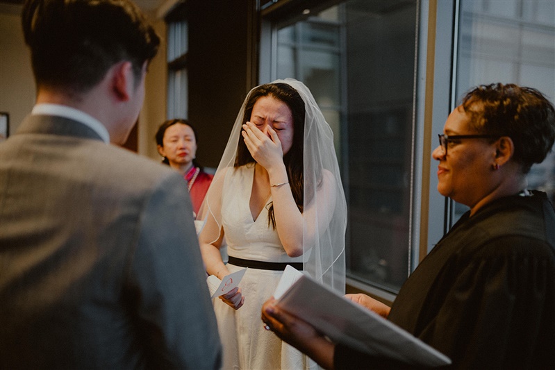 A bride holds her hand over her mouth as she cries tears of joy during her wedding ceremony. The foreground has the groom with his back to the camera and the officiant holding her ceremonial speech. The bride is wearing a white wedding dress with a black belt and long veil  pulled backwards.  The officiant is in a black gown.