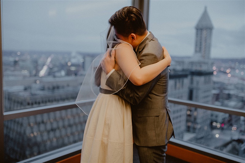A bride and groom embrace in a hug together after they have become officially married at the Seattle courthouse. The bride is wearing a white wedding dress with a black belt and view pulled back, while the groom is in a grey suit. The pair are standing in front of a window that overlooks Seattle.