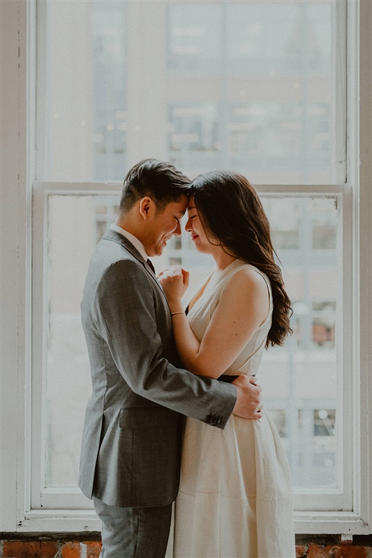 A bride and groom stand in front of a window and cuddle. The groom has his arms wrapped around the brides waist while both their foreheads lean in to touch. The groom is in a grey suit and the bride in a white wedding dress.