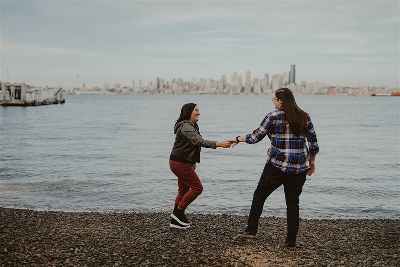 Two happy engaged women hold hands in front of the waterfront and cityscape for Seattle engagement photographer Chelsea Abril.