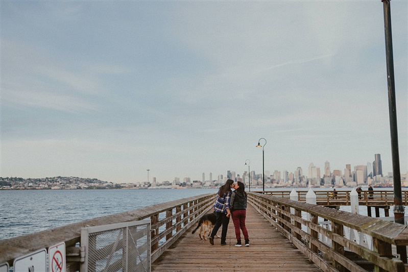 Two women kiss on the Alki Water Taxi Dock with their dog as they have their Seattle engagement photos taken.