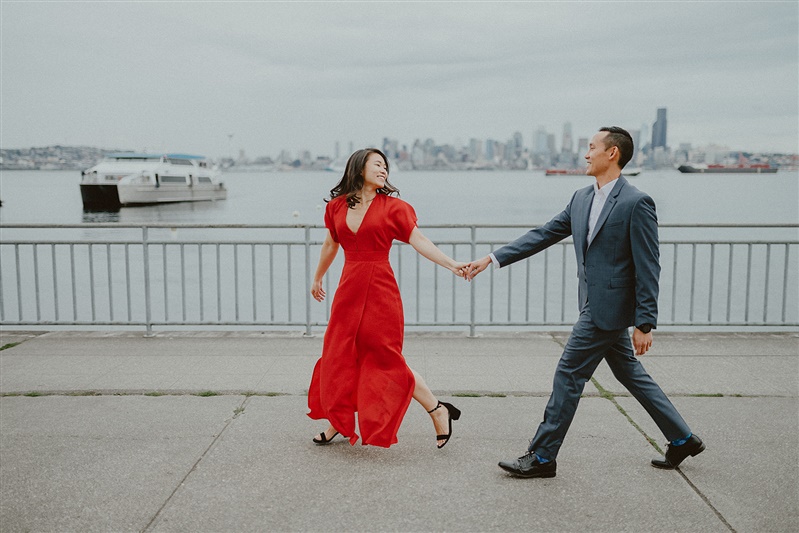 A woman in a red dress holds hands while walking with a man in a suit for their Seattle engagement photos at the Alki Water Taxi Dock.