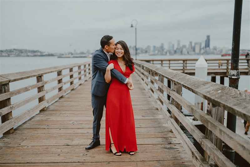 A man in a suit wraps his arms around his fiance wearing a long red dress, posing for engagement photos by the waterfront in Seattle