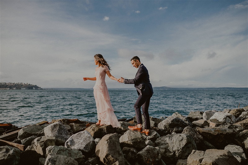 A woman in a pink dress holds hands with her fiance in a gray suit for engagement photos at the waterfront near Olympic Sculpture Park in Seattle.