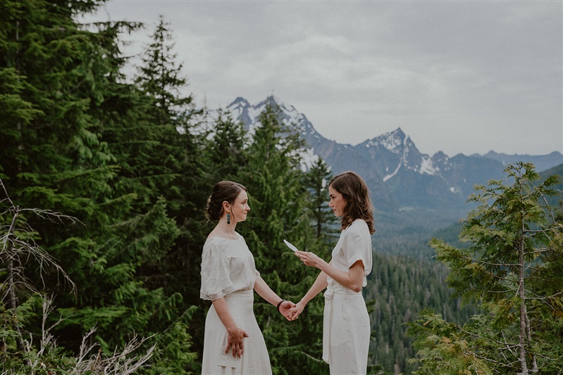 Two brides stand atop a mountain in Granite Falls and read their vows while Seattle elopemenent photographer Chelsea Anril captures the moment.