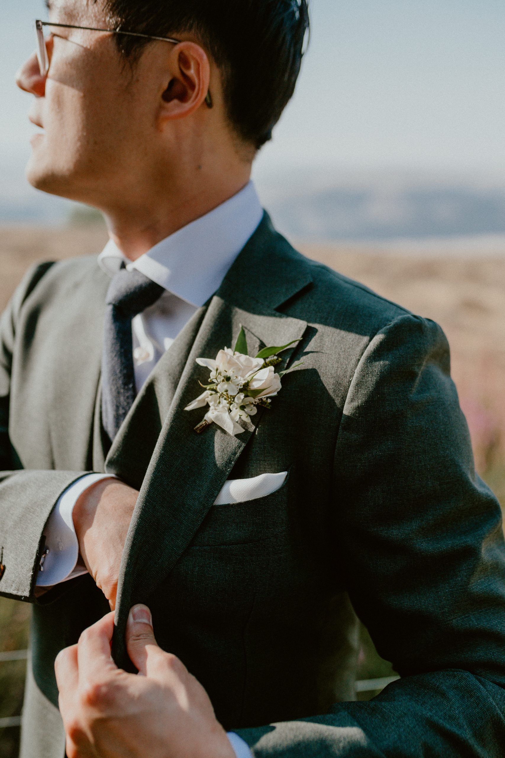 Groom with glasses dressed up fully in his wedding suit and looking out on to a field while putting something into his pocket while boutonniere pinned to the lapel of the grey wedding suit | Groom style tips, Groom wedding day inspiration, Groom Tips Wedding, Groom tips for men, Groom tips | chelseaabril.com 