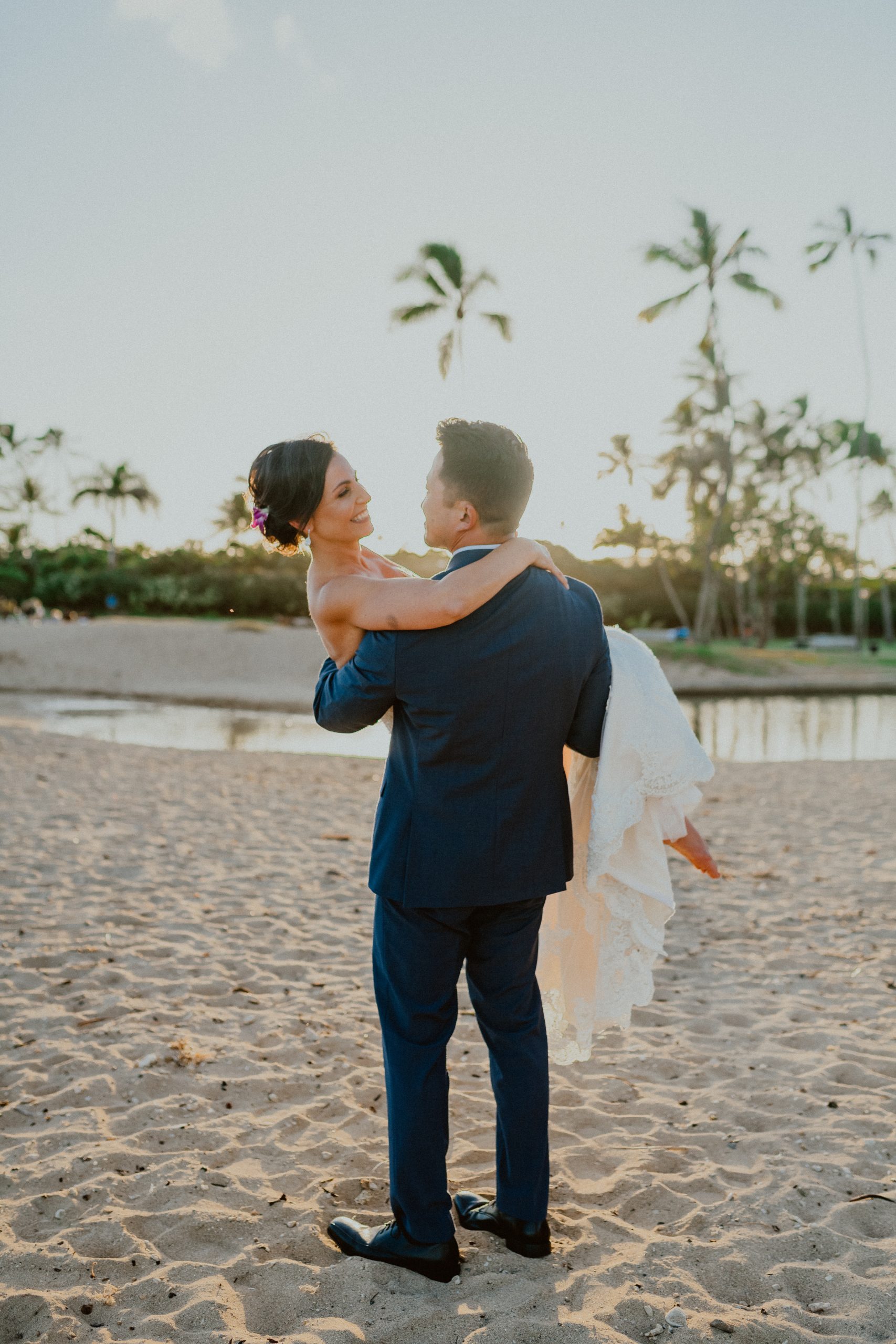 Groom in blue-grey suit style picks up bride as she smiles at him on the Oahu beach after their traditional Hawaii wedding ceremony | Oahu Wedding Photographer, Oahu Elopement Photographer, Destination Wedding Photographer, Destination Elopement Photographer, Newlywed moments ideas, Newlywed Photography inspiration, Hawaii Elopement ideas | chelseaabril.com