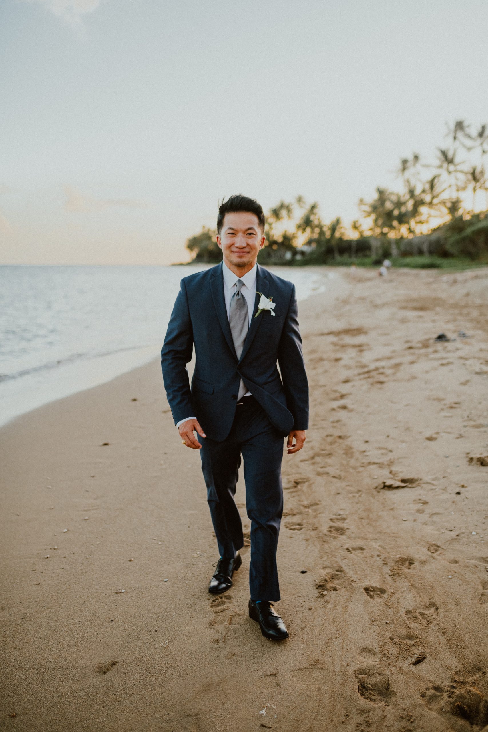 Groom walks towards Hawaii Wedding photographer on the Oahu beach while wearing his dark blue suit style with his white lily boutonniere on his lapel and black dress shoes for the Hawaii Wedding ceremony 